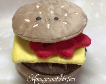 May include: A plush toy shaped like a hamburger with a smiling face. The hamburger is made of brown, yellow, and red plush fabric.