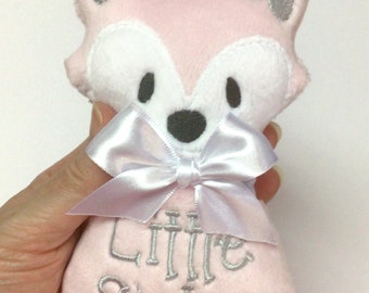 May include: A pink plush fox-shaped toy with white accents and a white satin bow. The text "Little Sister" is embroidered in gray on the toy. The fox has gray ears and black eyes. The toy is soft and cuddly.