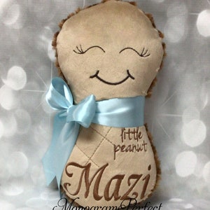 Puede incluir: Un peluche de color tostado con forma de cacahuete y un rostro bordado en marr&oacute;n con ojos cerrados y una sonrisa. El juguete tiene una cinta de sat&eacute;n azul claro atada alrededor y est&aacute; bordado con el texto little peanut y el nombre Mazi.