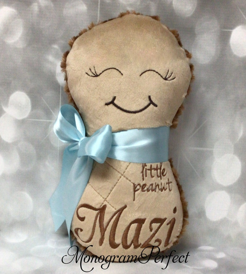Puede incluir: Un peluche de color tostado con forma de cacahuete y un rostro bordado en marr&oacute;n con ojos cerrados y una sonrisa. El juguete tiene una cinta de sat&eacute;n azul claro atada alrededor y est&aacute; bordado con el texto little peanut y el nombre Mazi.