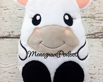 May include: White plush cow with black hooves, black eyes, and a pink nose. The cow is smiling and has a black embroidered line around its body. The text "Monogram Perfect" is embroidered on the cow's belly.
