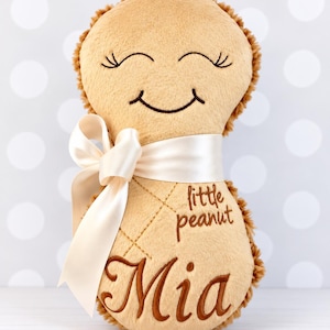 Puede incluir: Un peluche de color tostado con forma de cacahuete y textura suave. Presenta una cara sonriente bordada con los ojos cerrados, el texto "little peanut" y el nombre "Mia" en hilo marr&oacute;n. Una cinta de sat&eacute;n color crema est&aacute; atada en el centro.
