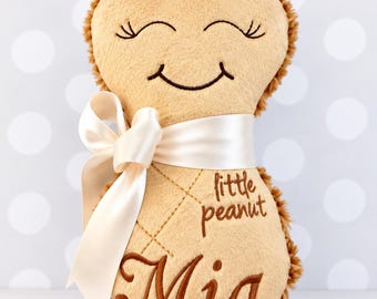 May include: A tan plush toy shaped like a peanut with a soft texture. It features an embroidered smiling face with closed eyes, the text "little peanut" and the name "Mia" in brown thread. A cream-colored satin ribbon is tied around the middle.