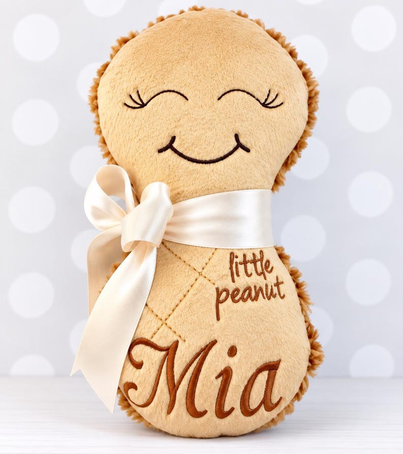 Puede incluir: Un peluche de color tostado con forma de cacahuete y textura suave. Presenta una cara sonriente bordada con los ojos cerrados, el texto "little peanut" y el nombre "Mia" en hilo marr&oacute;n. Una cinta de sat&eacute;n color crema est&aacute; atada en el centro.