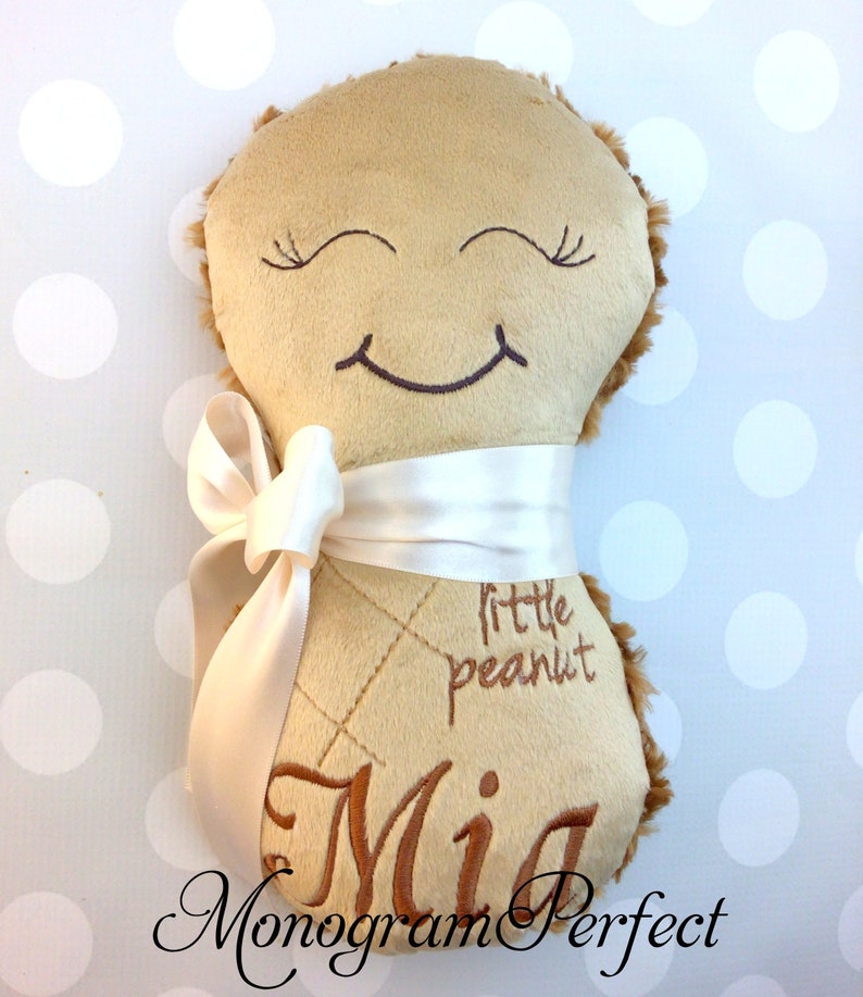 Puede incluir: Un peluche de color tostado con forma de cacahuete, con la parte delantera lisa y la trasera difusa. Presenta una cara sonriente bordada con los ojos cerrados, el texto "little peanut" y "Mia" en hilo marr&oacute;n y una cinta de sat&eacute;n blanca.