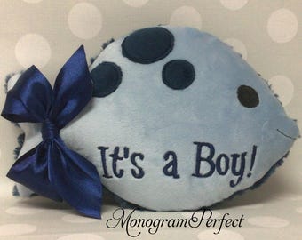 May include: A light blue plush fish-shaped pillow featuring dark blue circular spots, a dark blue satin ribbon bow attached to the tail, and the embroidered text "It's a Boy!" in dark blue thread. The text "MonogramPerfect" is printed below.