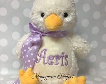 May include: A plush white duck toy with a soft, fuzzy texture, featuring a bright orange beak and feet. The duck wears a lavender ribbon with white polka dots tied around its neck. The name Aeris is embroidered in purple on its chest.