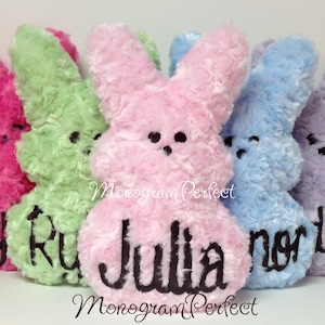 Puede incluir: Cinco juguetes de peluche con forma de conejito en tonos rosa, verde, rosa claro, azul y morado. Cada conejito tiene un nombre bordado en negro. El texto "Monogram Perfect" está escrito en cursiva en la parte inferior.