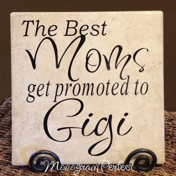 May include: A white ceramic tile with black text that reads "The Best Moms get promoted to Gigi". The tile is on a black metal stand.