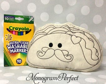 May include: A beige taco-shaped pillow with a black outline of a face, mustache, and heart. Next to it is a box of Crayola Ultra-Clean Washable Markers, 10 Classic Colors.