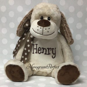 May include: A plush dog toy in cream and brown, with the name "Henry" embroidered on its chest. The dog has floppy ears, a brown nose, and a polka-dot ribbon. The words "Monogram Perfect" are also visible.