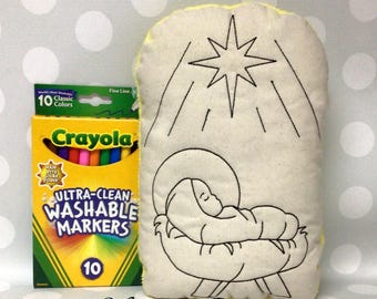 May include: A cream-colored, rectangular pillow with a black embroidered nativity scene and a star. A box of Crayola Ultra-Clean Washable Markers in 10 classic colors is next to the pillow. The text "Monogram Perfect" is at the bottom.