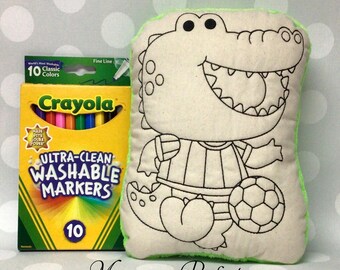 May include: A plush, cream-colored alligator-shaped pillow with black outline details, holding a soccer ball. The pillow is next to a box of Crayola Ultra-Clean Washable Markers, 10 classic colors. The text "Monogram Perfect" is at the bottom.