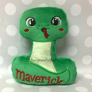 Op de afbeelding: Een groene pluche slang speelgoed met geborduurde details. De slang heeft grote, vriendelijke ogen, rode wangen en een rode tong die uitsteekt. De naam "Maverick" is rood op het lichaam geborduurd. Het speelgoed is ontworpen met een zachte, pluche textuur.