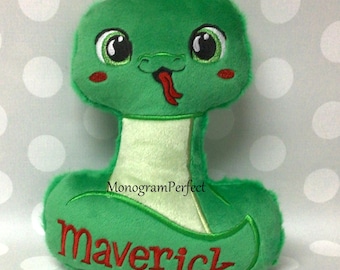May include: A green plush snake toy with embroidered details. The snake has large, friendly eyes, red cheeks, and a red tongue sticking out. The name "Maverick" is embroidered in red on the body. The toy is designed with a soft, plush texture.