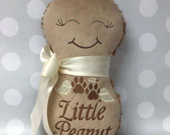 May include: A tan, peanut-shaped plush toy with a smooth front and fuzzy back. It features an embroidered smiling face, two brown paw prints with angel wings, and the text Little Peanut. A cream-colored satin ribbon is tied around the middle.
