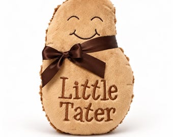 May include: A tan plush potato-shaped toy with a soft texture, featuring an embroidered smiling face and the text Little Tater in brown thread. A dark brown satin ribbon is tied in a bow around the upper portion of the plush object.