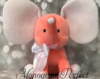 May include: A plush peach-colored elephant toy with large white ears and a white bow. The elephant has a pink nose and a white spot on its forehead.