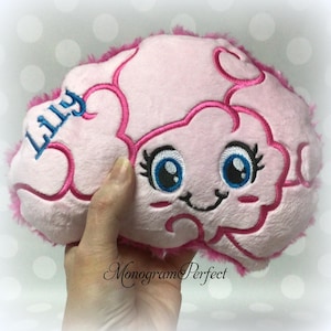 Peut inclure: Un coussin rose en forme de cerveau avec des détails brodés. Le coussin présente un visage souriant avec des yeux bleus et des cils. Le nom "Lilly" est brodé en bleu sur le côté. Le coussin a une bordure rose duveteuse.