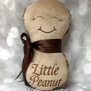 May include: A plush peanut-shaped toy with a smiling face and embroidered eyelashes. The toy is brown and has a brown satin ribbon tied around it. The text "Little Peanut" is embroidered on the front of the toy.