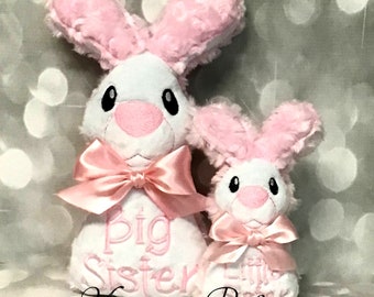 May include: Two plush pink and white bunny toys. The larger bunny has a pink bow and says "Big Sister" in pink embroidery. The smaller bunny has a pink bow and says "Little Sister" in pink embroidery.
