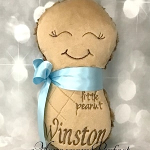 Puede incluir: Un juguete de peluche color canela con forma de cacahuete y textura suave, con ojos cerrados y boca sonriente bordados. El juguete tiene una cinta de sat&eacute;n azul claro atada alrededor de la parte central y est&aacute; bordado con el texto little peanut y Winston.
