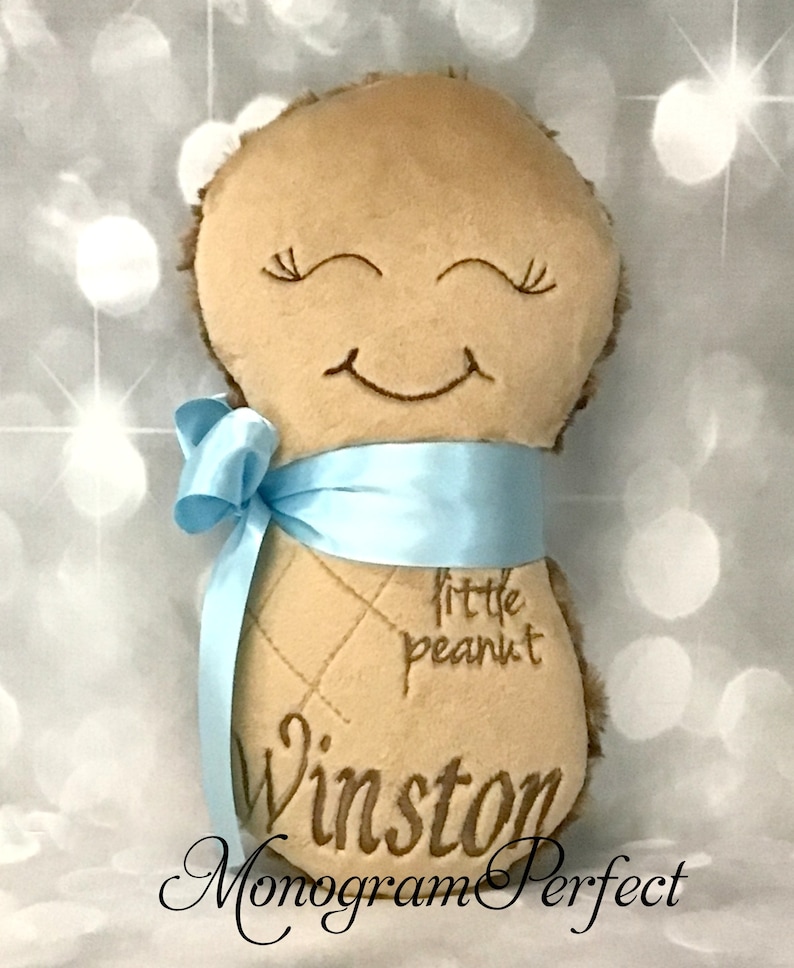 Puede incluir: Un juguete de peluche color canela con forma de cacahuete y textura suave, con ojos cerrados y boca sonriente bordados. El juguete tiene una cinta de sat&eacute;n azul claro atada alrededor de la parte central y est&aacute; bordado con el texto little peanut y Winston.