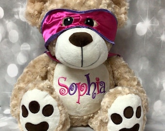 May include: A beige plush teddy bear wearing a pink and purple superhero mask. The bear has brown paw pads and is embroidered with the name "Sophia".