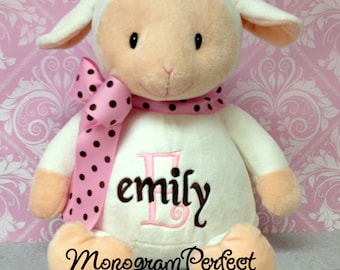 May include: White plush lamb with a pink and black polka dot ribbon around its neck. The lamb is embroidered with the name "emily" in brown and pink thread. The lamb is sitting on a pink and white patterned background. The text "Monogram Perfect" is embroidered on the lamb's belly.