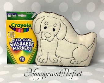 May include: A beige dog-shaped pillow with black embroidered outlines, next to a box of Crayola Ultra-Clean Washable Markers in 10 classic colors. The text "Monogram Perfect" is at the bottom.