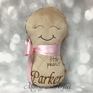 Puede incluir: Un peluche de color tostado con forma de cacahuete, con ojos cerrados y boca sonriente bordados. Presenta una cinta de sat&eacute;n rosa claro atada alrededor del centro, con el texto "little peanut" y el nombre "Parker" bordados en hilo marr&oacute;n al frente.