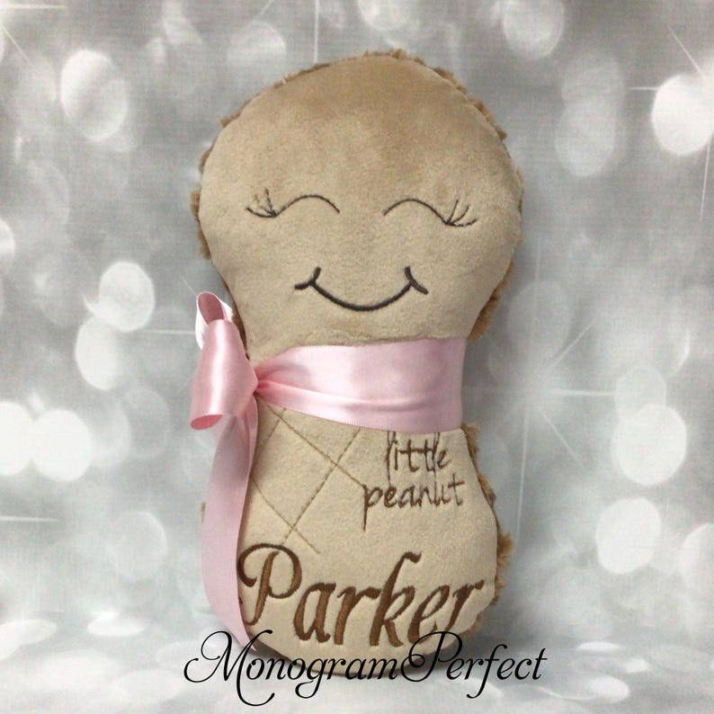 Puede incluir: Un peluche de color tostado con forma de cacahuete, con ojos cerrados y boca sonriente bordados. Presenta una cinta de sat&eacute;n rosa claro atada alrededor del centro, con el texto "little peanut" y el nombre "Parker" bordados en hilo marr&oacute;n al frente.