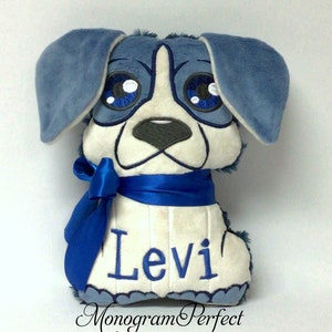 Op de afbeelding: Blauw en wit pluche hond met grote ogen en een blauw lint om zijn nek. De hond is geborduurd met de naam "Levi".