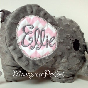 Puede incluir: Elefante de peluche gris con un parche de tela de chevron rosa y blanco en la cabeza. El parche tiene el nombre "Ellie" bordado. El elefante tiene una cola de satén gris.