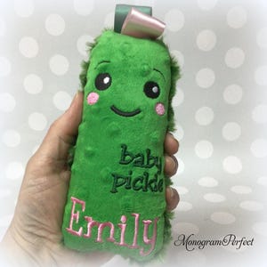 May include: A green plush baby pickle toy with embroidered details. The toy features a smiling face with pink cheeks, the words "baby pickle" and the name "Emily" embroidered in pink. A green ribbon and pink bow are attached at the top.