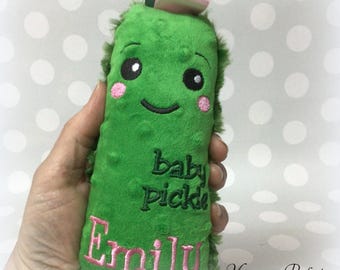 May include: A plush green baby pickle toy with embroidered details. The toy has a smiling face with pink cheeks, the words "baby pickle" and the name "Emily" embroidered in pink. A green ribbon and pink bow are attached at the top.