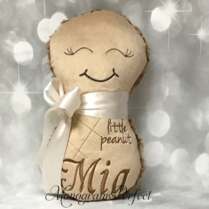 Puede incluir: Un juguete de peluche color canela con forma de cacahuete y textura suave, con ojos cerrados y una boca sonriente bordados. El juguete est&aacute; envuelto con una cinta de sat&eacute;n blanca atada en un lazo y muestra el texto bordado "little peanut" y "Mia".
