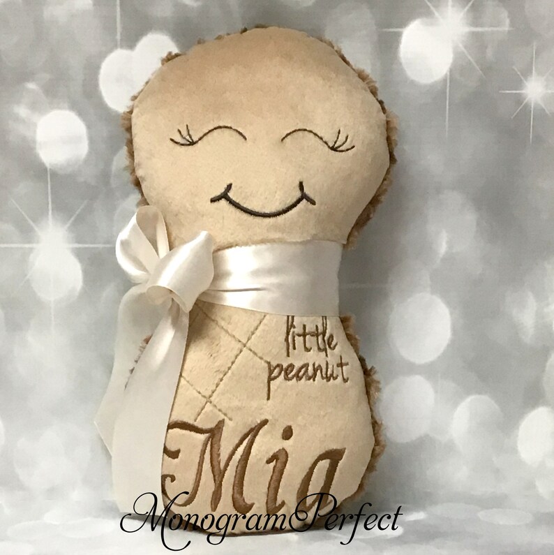 Puede incluir: Un juguete de peluche color canela con forma de cacahuete y textura suave, con ojos cerrados y una boca sonriente bordados. El juguete est&aacute; envuelto con una cinta de sat&eacute;n blanca atada en un lazo y muestra el texto bordado "little peanut" y "Mia".
