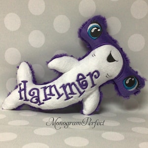 May include: A white and purple plush hammerhead shark toy. The word "Hammer" is embroidered in purple on the shark's body. The shark has large, blue-eyed eyes and a soft, fuzzy texture. The text "Monogram Perfect" is below the shark.