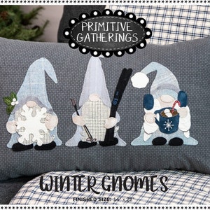 May include: A gray pillow with three winter gnomes embroidered on it. The gnomes are wearing blue hats and holding a snowflake, skis, and a mug of hot chocolate. The pillow has the text "Primitive Gatherings" and "Winter Gnomes" on it.