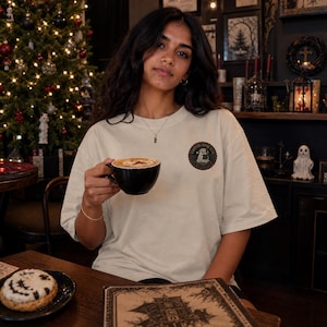 May include: A beige short-sleeved t-shirt featuring a circular graphic on the left chest. The graphic depicts a ghost holding a coffee cup with the text "COFFEE SOCIAL CLUB" written in a circular border around the central illustration.