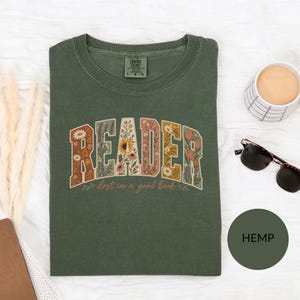 Pode incluir: Uma t-shirt verde-oliva com a palavra READER em letras grandes de bloco preenchidas com padrões florais, com o texto lost in a good book por baixo. A t-shirt tem uma etiqueta Comfort Colors no pescoço e é exibida com óculos de sol.