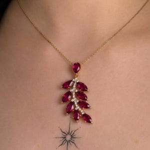 May include: A gold-tone necklace featuring a pendant shaped like a leafy branch. The pendant is adorned with multiple marquise-cut deep red gemstones and small, clear, round-cut accent stones set along the central stem of the branch design.