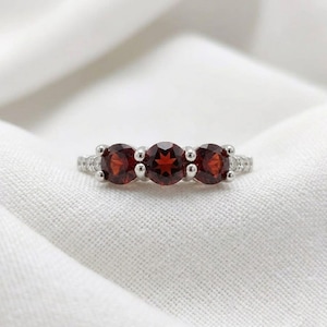 Könnte beinhalten: Ein silberfarbener Ring mit drei runden, tiefroten Granat-Edelsteinen in horizontaler Anordnung. Die Ringschiene ist an den Seiten mit kleinen, klaren Pavé-gefassten Steinen besetzt, was ein klassisches Drei-Stein-Design mit poliertem Finish ergibt.