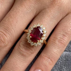 May include: A gold ring featuring a large, oval-cut deep red gemstone at the centre, surrounded by a halo of small, round, clear white diamonds. The band is crafted from polished yellow gold, creating a classic and elegant jewellery piece.