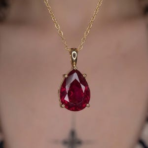 Può includere: Un ciondolo con pietra preziosa rosso intenso a forma di pera, montato su una montatura in metallo dorato e sospeso a una delicata catenina dorata. La pietra sfaccettata riflette la luce, mettendo in risalto la sua tonalità cremisi.