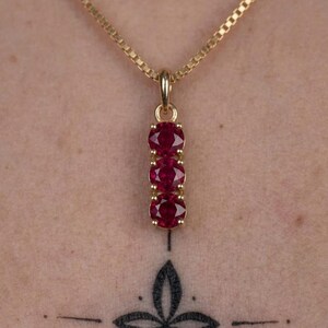 May include: A gold pendant necklace featuring three round, deep red rubies set vertically in a gold frame. The pendant hangs from a delicate gold box chain. The stones are arranged in a straight line, each secured by four small gold prongs.