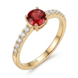 Könnte beinhalten: Ein goldfarbener Ring mit einem runden, facettierten roten Mittelstein in einer Vier-Krappen-Fassung. Die Ringschiene ist im oberen Bereich mit einer Reihe kleiner, klarer, rund geschliffener Steine besetzt und trägt innen die Prägung 925.