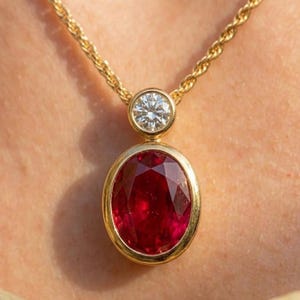 May include: A gold chain necklace featuring a pendant with a large, oval-cut, deep red gemstone set in a smooth gold bezel. Above the oval stone sits a smaller, round-cut, clear white gemstone in a matching gold bezel setting, creating a two-tier design.