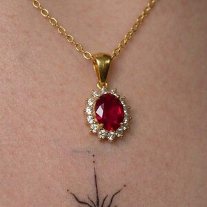 Peut inclure: Un collier à chaîne dorée avec un pendentif en pierre précieuse rouge foncé de taille ovale, entouré d'un halo de petites pierres claires de taille ronde. Le pendentif est suspendu à une fine chaîne à maillons dorés sur la peau.